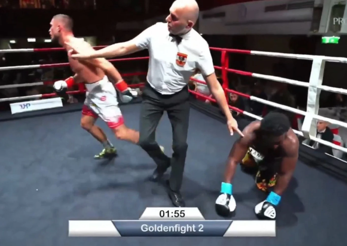 Sędzia oddziela dwóch zawodników w ringu bokserskim. Jeden z bokserów klęczy na macie po ciosie, drugi odchodzi w stronę narożnika. Na dole ekranu widoczny jest zegar i nazwa wydarzenia Goldenfight 2.