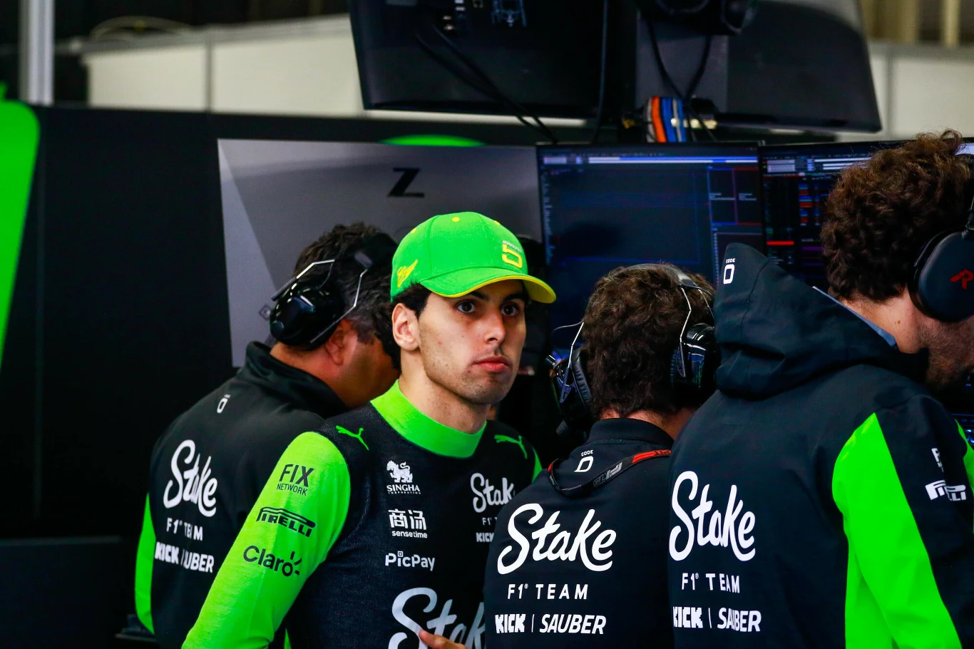 Młody mężczyzna w zielonej czapce i kombinezonie teamu wyścigowego Stake F1 Team Nick Sauber stoi w pobliżu technicznego zaplecza, otoczony przez członków zespołu skupionych na monitorach.