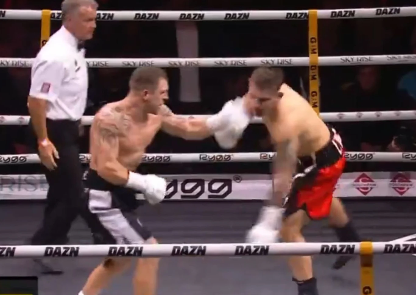 Dwóch bokserów w trakcie pojedynku na ringu, jeden z nich właśnie zadaje cios w stronę drugiego, a pomiędzy nimi stoi sędzia obserwujący przebieg walki. Na tle widać ogrodzenie ringu z logo DAZN.