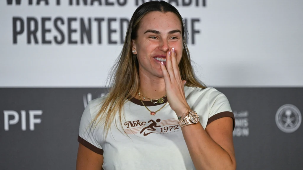 Aryna Sabalenka po finale WTA Finals Kobieta ubrana w jasną koszulkę z logo Nike, uśmiecha się i zasłania usta dłonią, w tle rozmazany napis i logotypy.