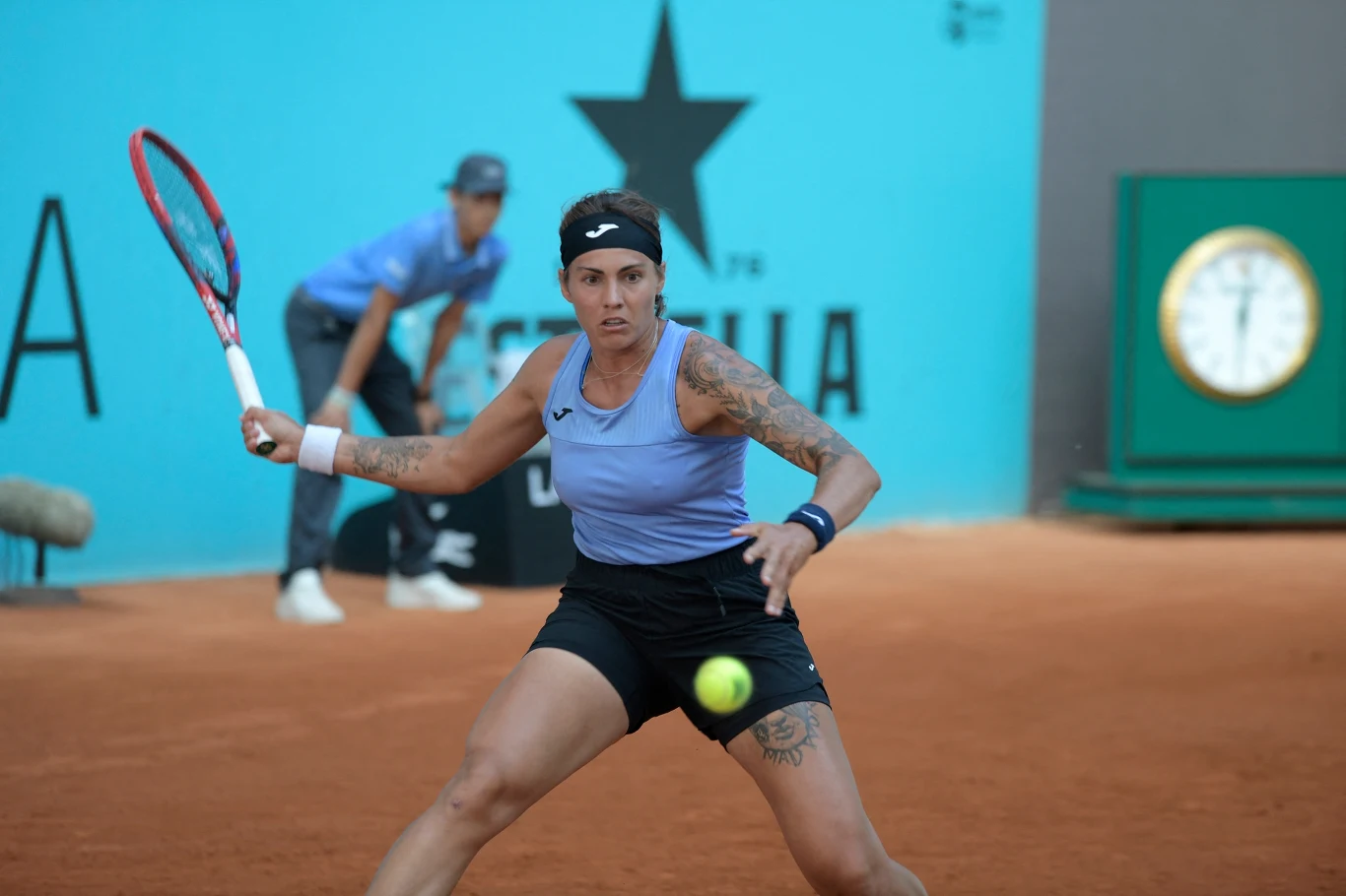 Tenisistka w sportowym stroju i z tatuażami przygotowuje się do odbioru piłki na korcie ziemnym, w tle sędzia i zegar turniejowy.