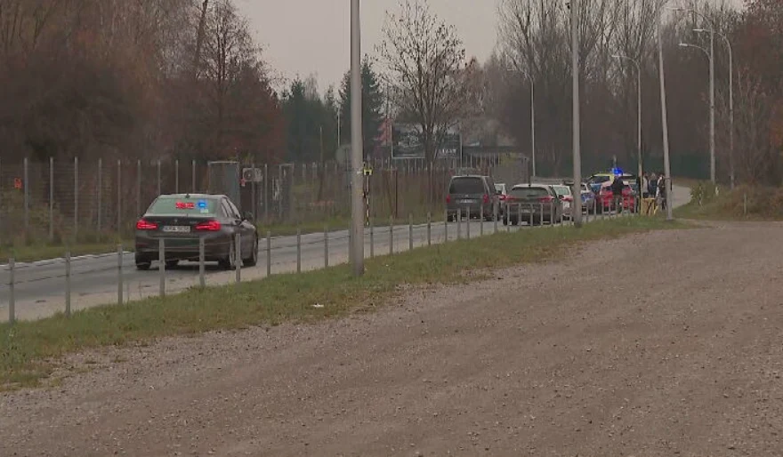 Kolumna samochodów, w tym radiowóz z niebieskimi światłami, jedzie wzdłuż wąskiej, ogrodzonej drogi, wokół znajdują się drzewa bez liści i pole.