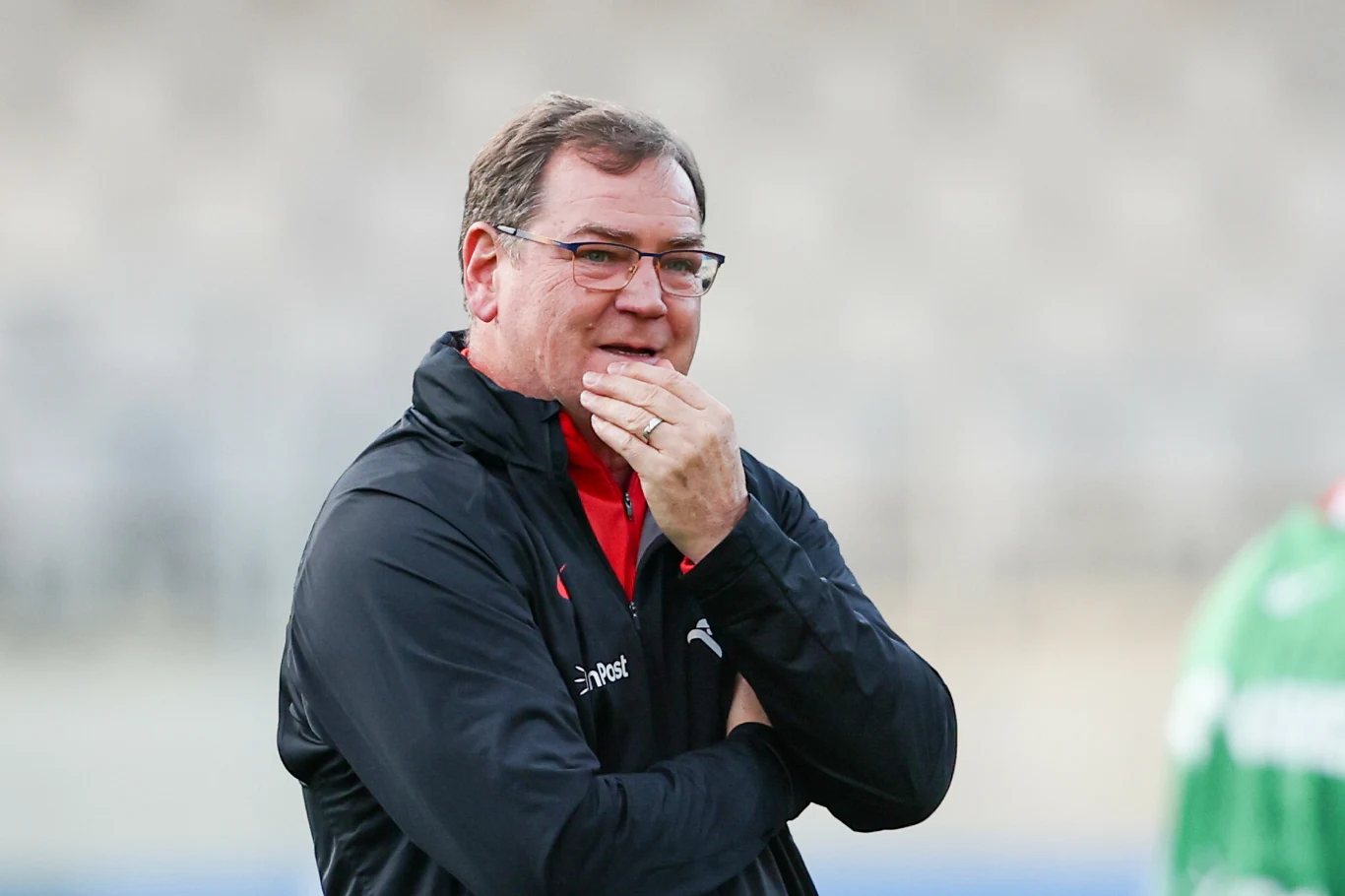 Mężczyzna w sportowej kurtce z zamyślonym wyrazem twarzy, opiera dłoń na brodzie, spogląda w dal. Tło rozmyte, w prawym rogu widoczna sylwetka osoby w zielonej kurtce.