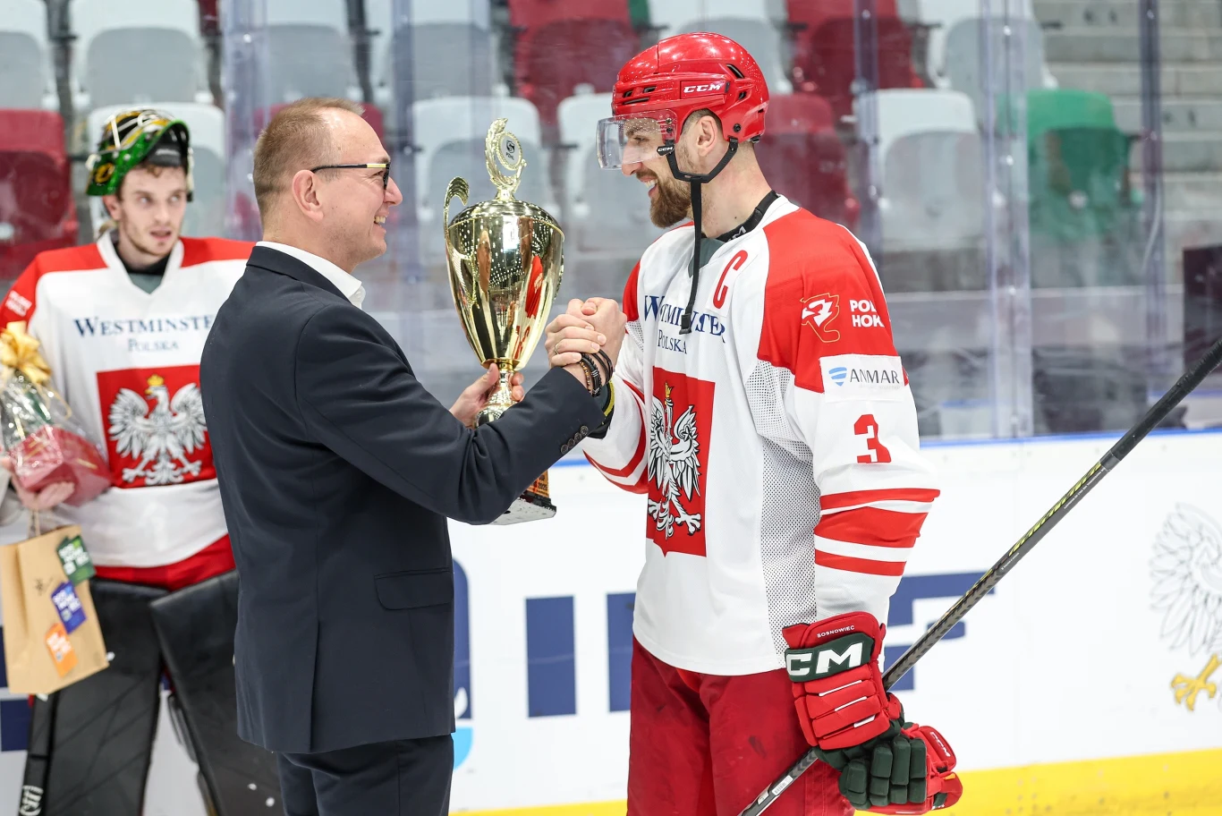 Bartosz Ciura odbiera puchar za zwycięstwo w turnieju od Krzysztofa Woźniaka Kapitan drużyny hokejowej w biało-czerwonym stroju odbiera puchar od elegancko ubranego mężczyzny. W tle bramkarz z bukietem kwiatów w ręku obserwuje sytuację. Wszyscy znajdują się na lodowisku, a trybuny są częściowo puste.