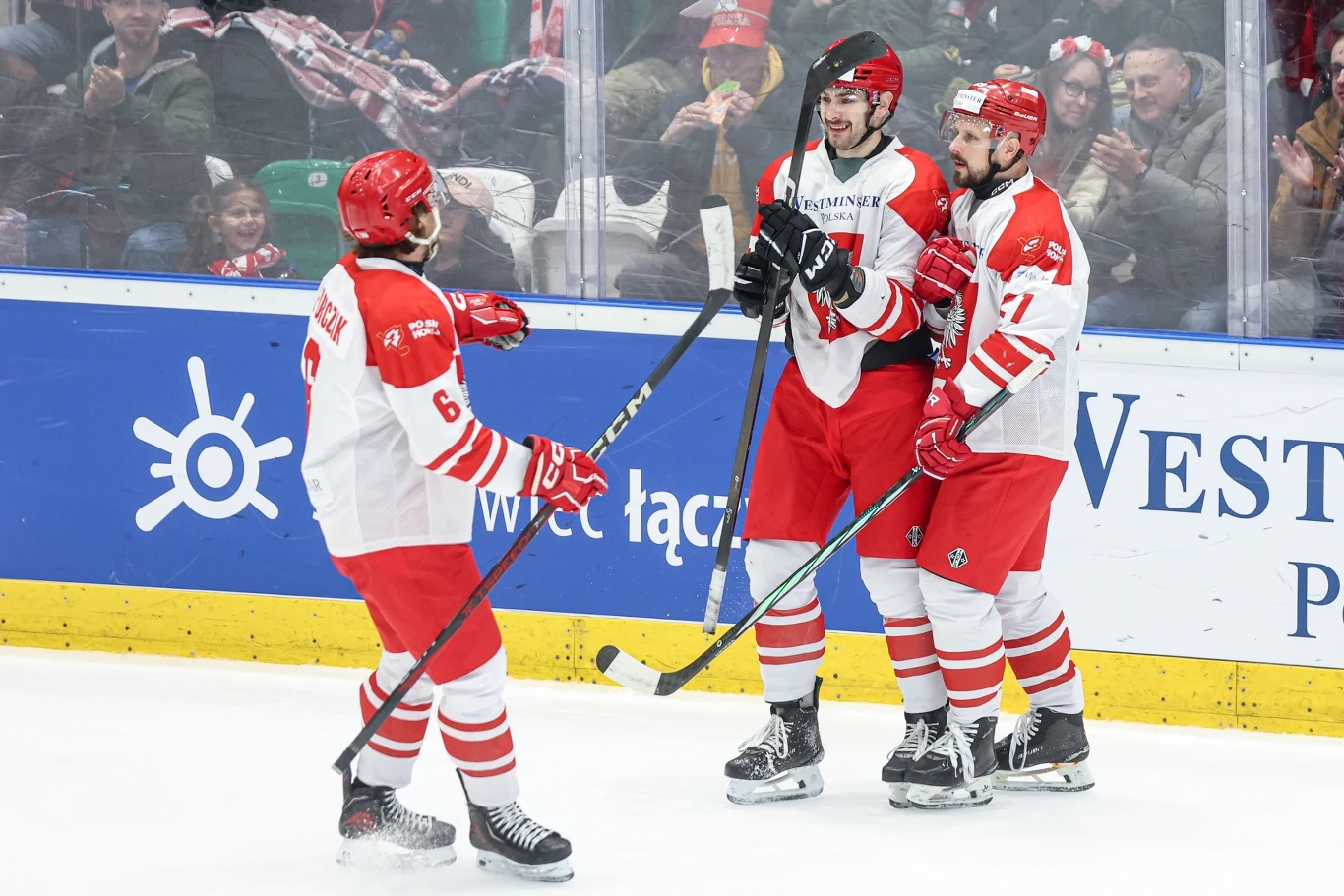 Radość polskich hokeistów Trzech hokeistów w biało-czerwonych strojach świętuje zdobycie bramki na lodowisku, w tle widownia kibicująca zespołowi.