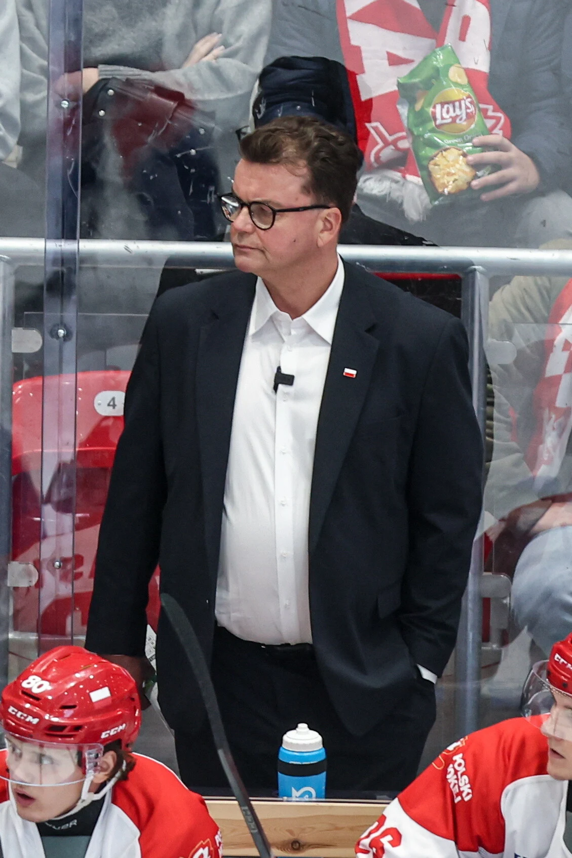 Pekka Tirkkonen Mężczyzna w garniturze stoi przy bandzie lodowiska, za nim kibice, dwóch hokeistów drużyny w czerwono-białych strojach siedzi na ławce obok niebieskiego bidonu, w tle osoby z szalami i paczką chipsów.