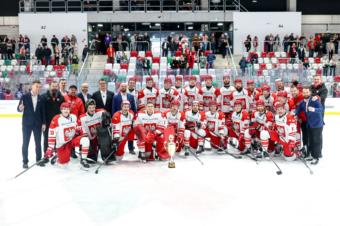 Reprezentacja Polski w hokeju na lodzie Grupa hokeistów ubranych w biało-czerwone stroje stoi na lodowisku wraz ze sztabem trenerskim i oficjelami. Na pierwszym planie znajduje się zdobyty puchar. W tle widoczna jest trybuna z kibicami.