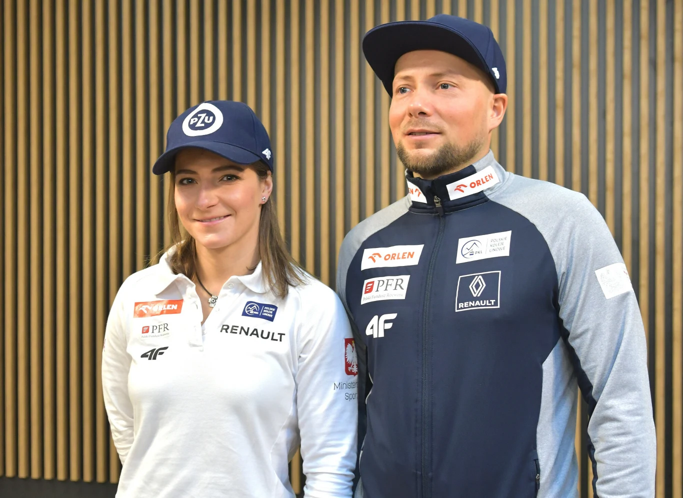 Dwie osoby ubrane w sportowe stroje z logotypami sponsorów, stoją obok siebie na tle ściany z pionowymi, drewnianymi panelami. Kobieta i mężczyzna mają na głowach czapki z daszkiem, oboje uśmiechnięci.