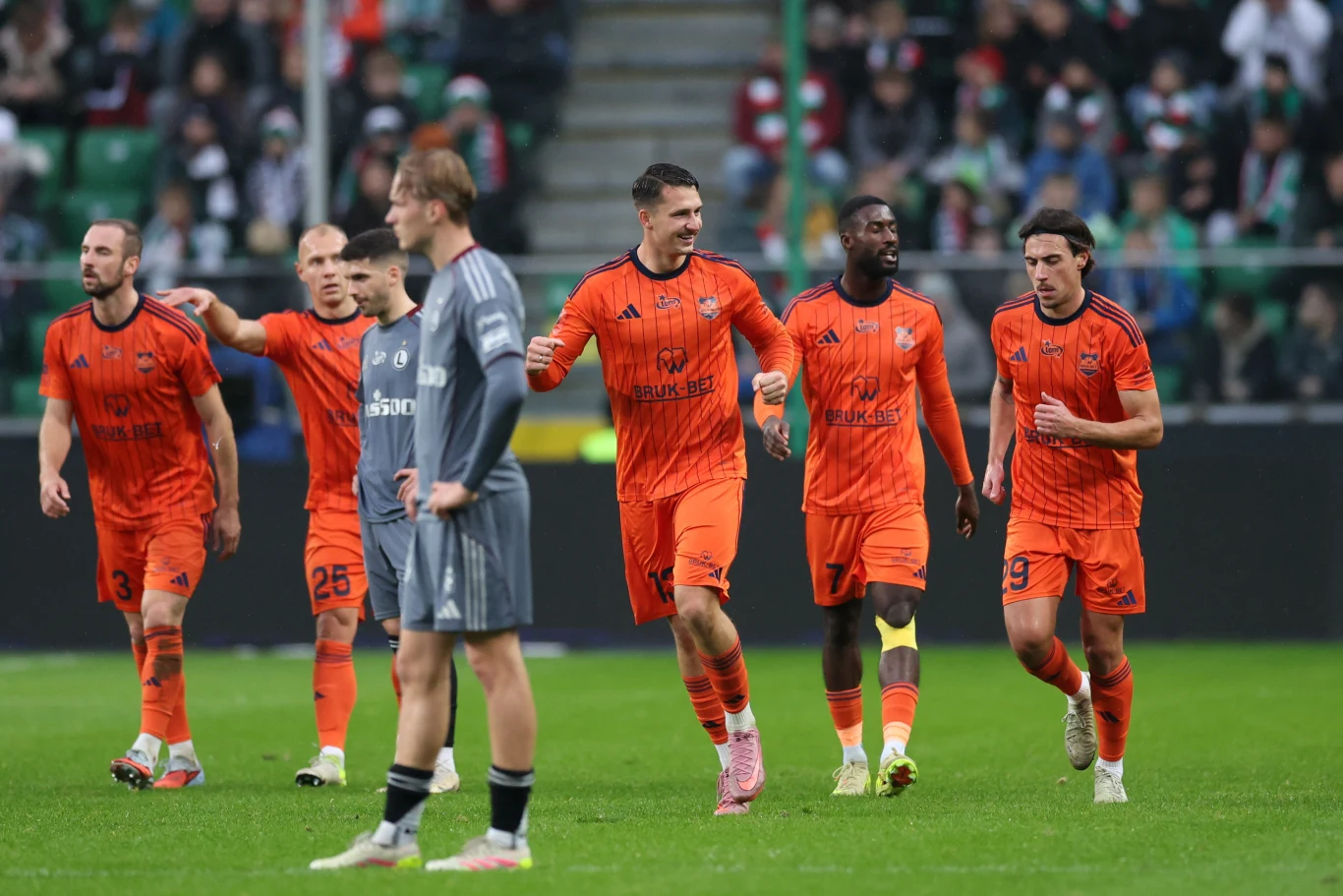 Legia Warszawa - Termalica Nieciecza Piłkarze w pomarańczowych strojach świętują zdobycie bramki na murawie stadionu, w tle zawodnik drużyny przeciwnej w szarym stroju oraz trybuny z kibicami.