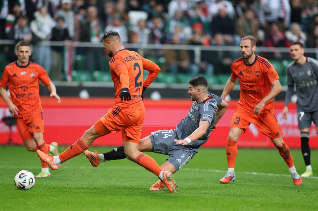 Dynamiczna akcja podczas meczu piłki nożnej, zawodnik w pomarańczowym stroju próbuje przejąć piłkę od przeciwnika w szarym stroju, na tle boiska oraz trybun z kibicami.