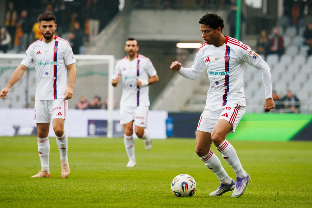 Świetny mecz w Kielcach pomiędzy Koroną, a Rakowem Piłkarz w białym stroju z piłką przy nodze podczas meczu piłkarskiego, w tle dwóch innych zawodników tej samej drużyny na zielonej murawie, trybuny z kibicami są w tle, atmosfera stadionowa.