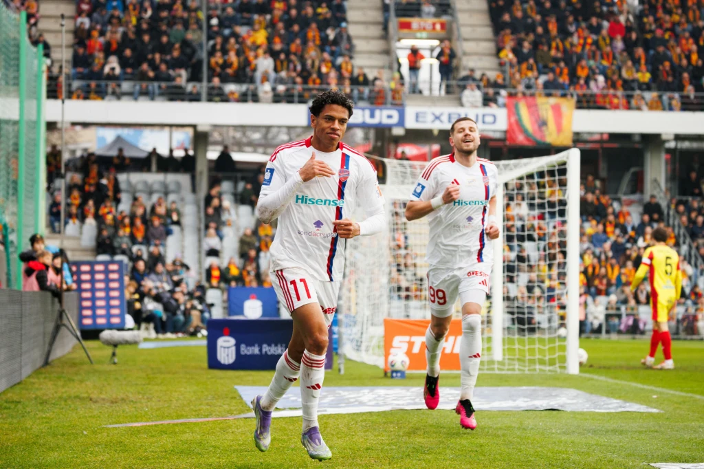 Adriano Amorim wyprowadził Raków na prowadzenie Dwóch piłkarzy w białych strojach z czerwonymi elementami biegnie obok siebie na tle bramki i pełnych trybun stadionu, w tle widoczna jest publiczność oraz zawodnicy przeciwnej drużyny w żółto-czerwonych strojach.