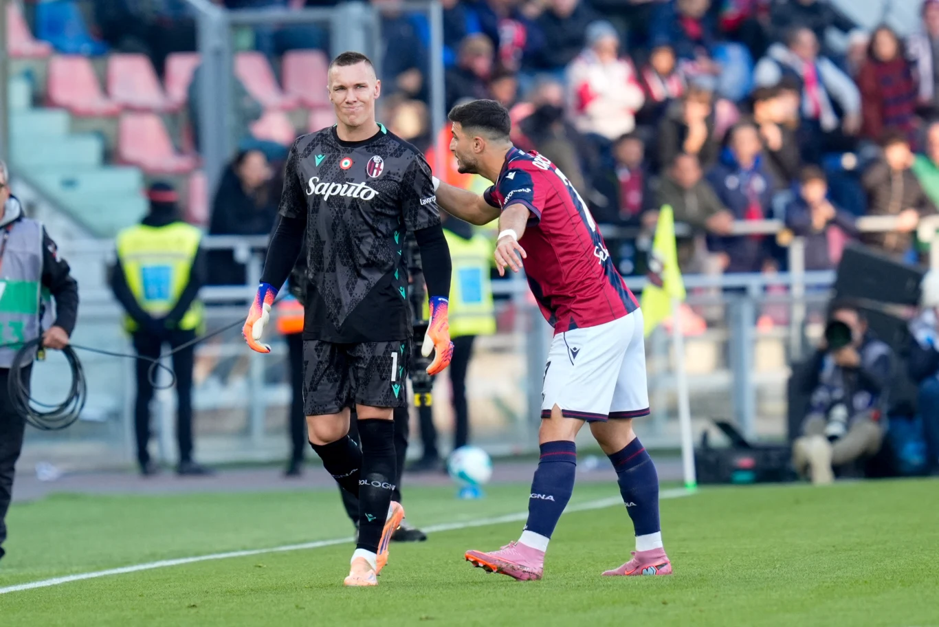 Dwóch piłkarzy na boisku podczas meczu, jeden ubrany na czarno jest bramkarzem, drugi w bordowym stroju jest zawodnikiem z pola. Za nimi widownia na stadionie, część osób w żółtych kamizelkach ochrania wydarzenie sportowe.