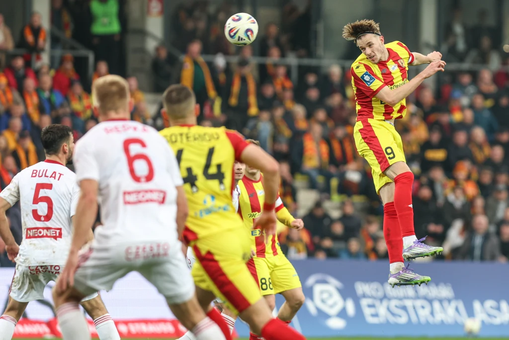 Korona Kielce - Raków Częstochowa Piłkarz w żółto-czerwonym stroju wyskakuje dynamicznie do piłki głową podczas meczu piłkarskiego, otoczony przez zawodników przeciwnika w białych strojach oraz kolegów z drużyny, na tle pełnych trybun.