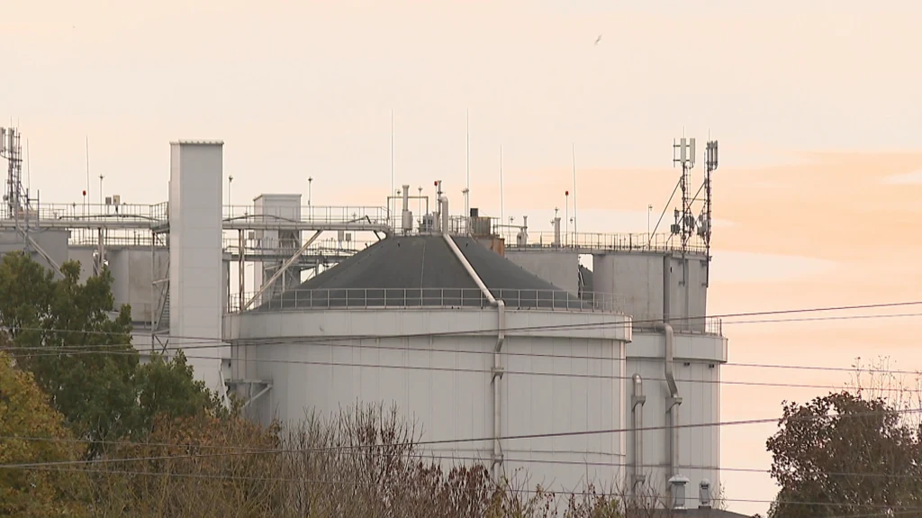 Duży, przemysłowy zbiornik na ciecz z antenami i instalacjami technicznymi na dachu, otoczony przez drzewa i linie energetyczne w tle nieba o ciepłej barwie.