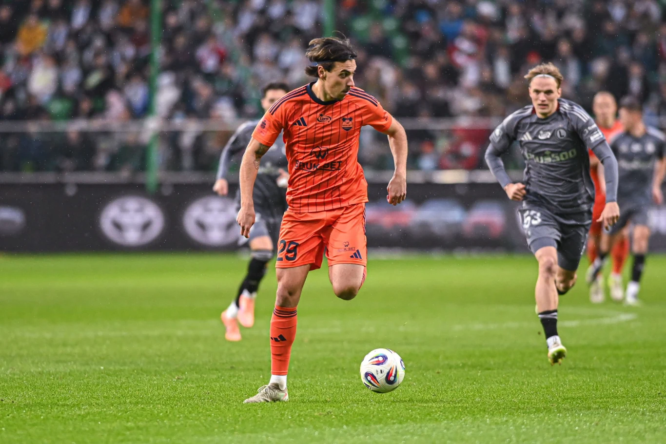 Legia Warszawa - Termalica Nieciecza Piłkarz w pomarańczowym stroju prowadzi piłkę po zielonej murawie podczas meczu piłkarskiego, w tle widać przeciwników w szarych strojach i zapełnione trybuny.