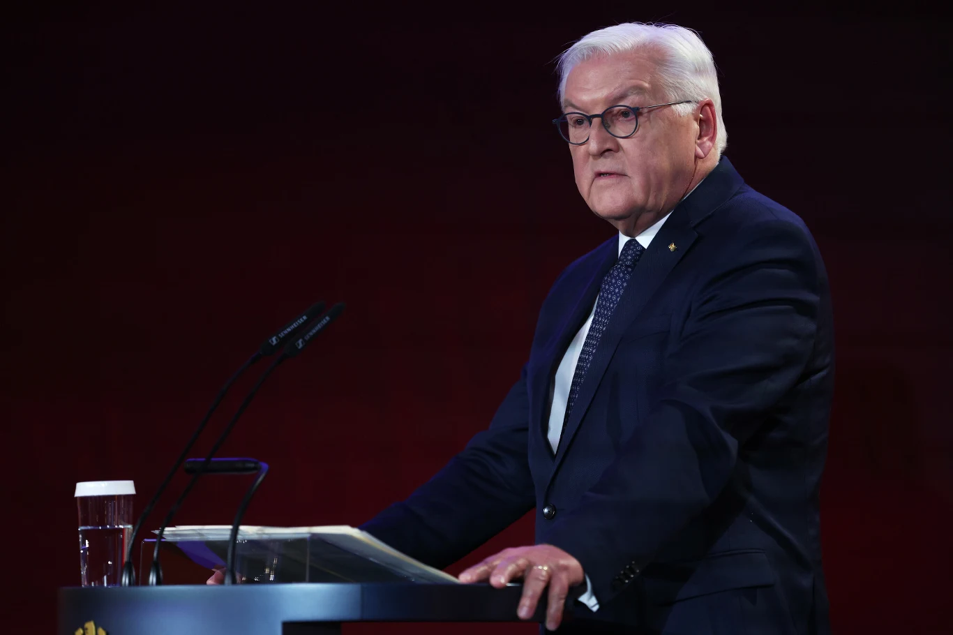 Prezydent Niemiec jest zdania, że demokracja w jego kraju znalazła się w trudnej sytuacji, a zagrażają jej m.in. ekstremiści Starszy mężczyzna w okularach ubrany w garnitur przemawia za mównicą z mikrofonami, obok stoi szklanka z wodą. Tło ciemne, jednolite.