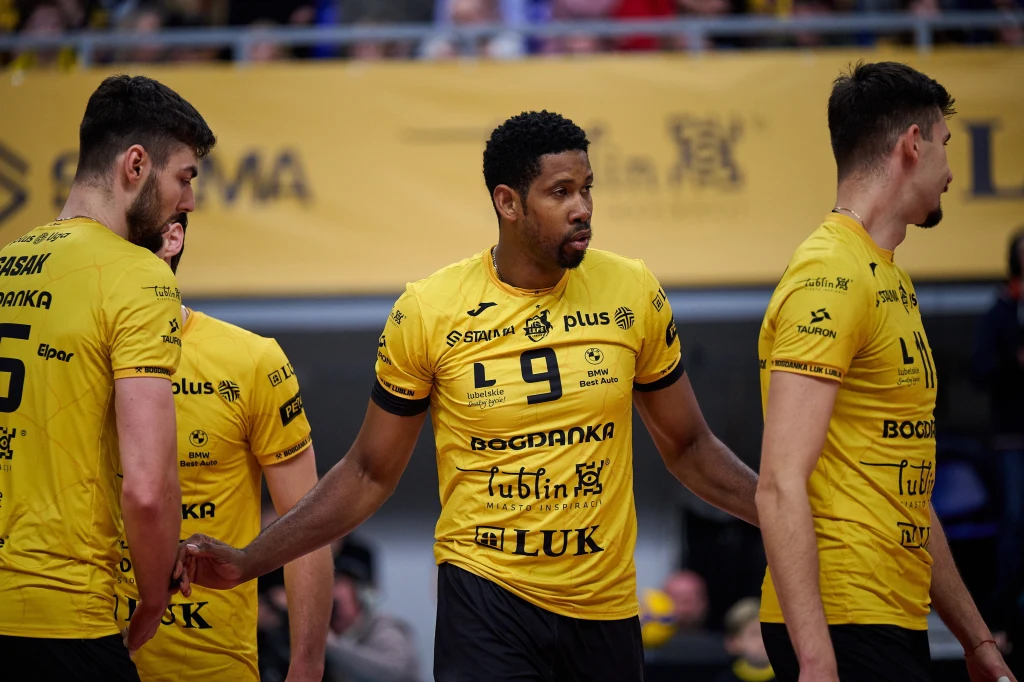 Wilfredo Leon Grupa siatkarzy w żółtych koszulkach drużyny LUK Lublin stoi na boisku w trakcie meczu, koncentrując się na rozmowie lub naradzie, w tle kibice i banery reklamowe.