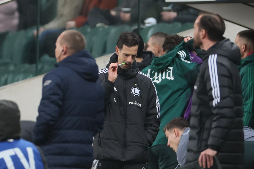 Inaki Astiz, aktualny trener Legii Warszawa Trener drużyny piłkarskiej ubrany w kurtkę z logo klubu analizuje sytuację na boisku, trzymając coś w dłoni, wokół niego stoją inni członkowie sztabu oraz zawodnicy rezerwowi.