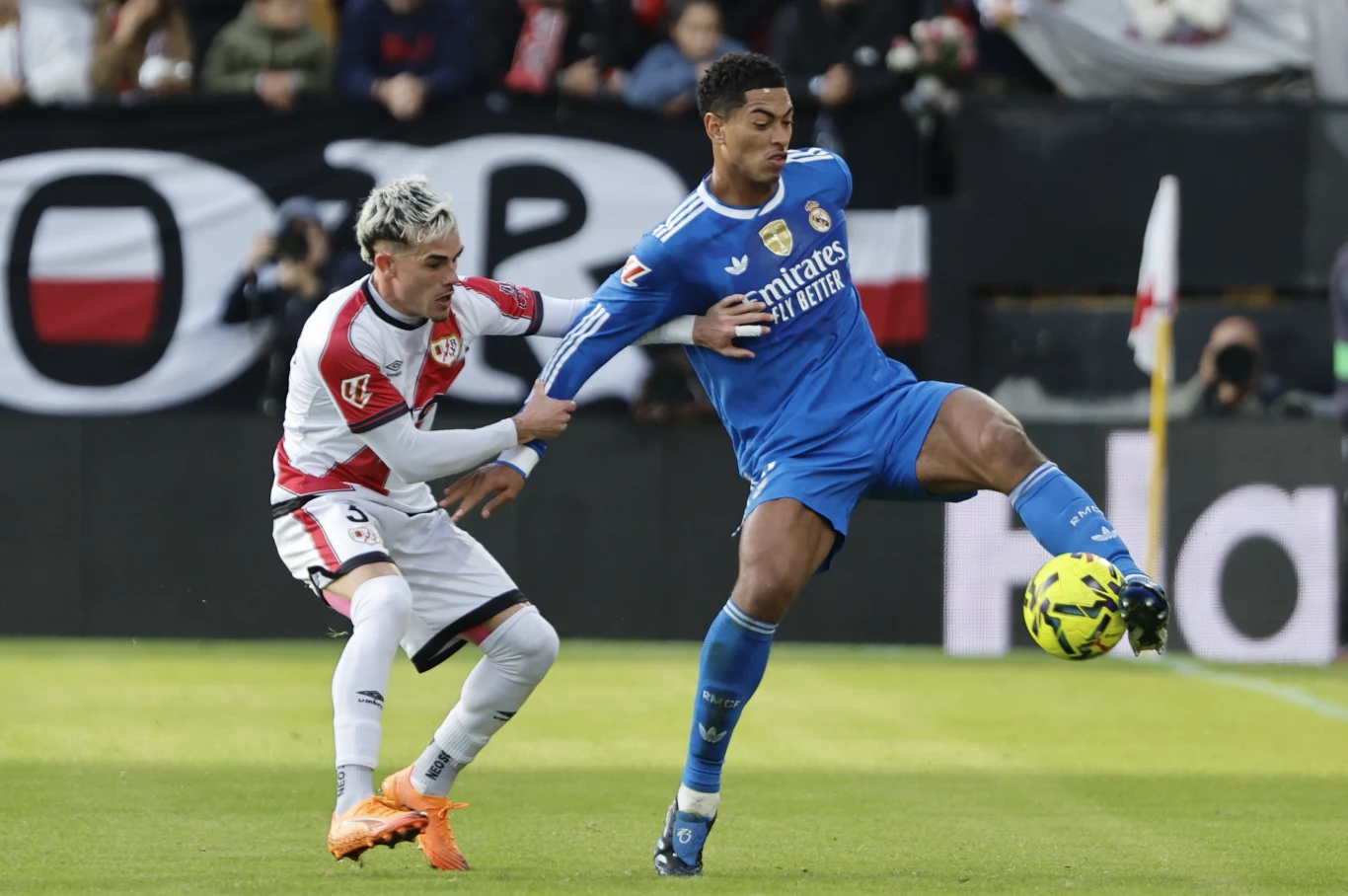 Dwóch piłkarzy w akcji podczas meczu, zawodnik w niebieskim stroju kontroluje piłkę, a przeciwnik w biało-czerwonym stroju próbuje go powstrzymać, w tle widoczni kibice oraz fragmenty stadionu.