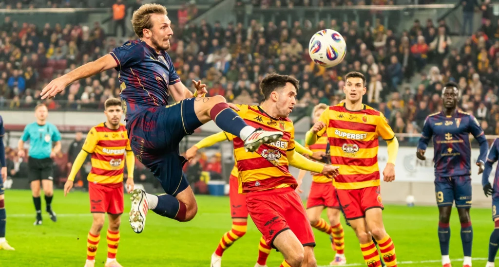 Piłkarz w granatowo-czerwonym stroju wyskakuje do piłki, próbując oddać strzał głową, podczas gdy zawodnik w żółto-czerwonym komplecie próbuje go powstrzymać. W tle widoczni są inni piłkarze oraz publiczność na stadionie.