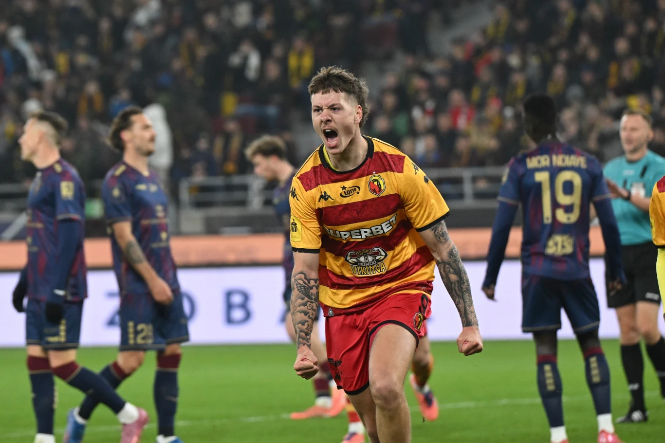 Piłkarz w żółto-czerwonym stroju wyraża silne emocje podczas meczu piłki nożnej, zaciśnięte pięści i twarz ukazują determinację. W tle pozostali zawodnicy oraz sędzia na murawie stadionu, tłum kibiców na trybunach.