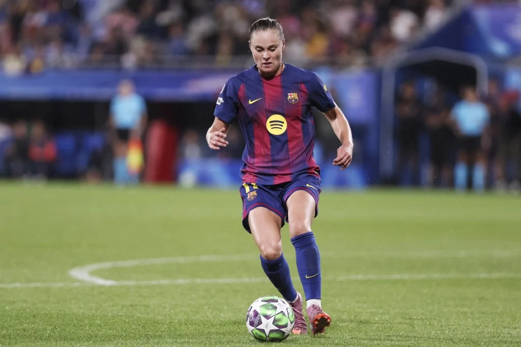 Piłkarka w barwach FC Barcelony prowadzi piłkę na boisku podczas meczu, skupiona na grze, w tle widoczni są sędziowie i rozmyci kibice.