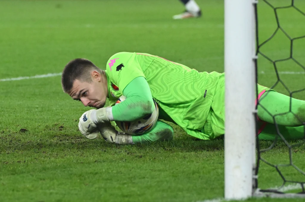Bramkarz w jasnozielonym stroju leży na murawie i mocno przytrzymuje piłkę przy linii bramkowej, skupiając się na obronie.