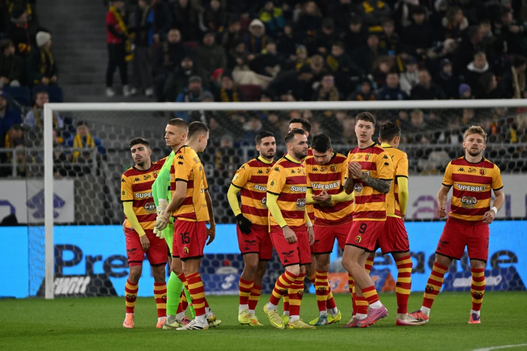 Grupa piłkarzy ubranych w żółto-czerwone stroje sportowe zgromadzona na boisku piłkarskim, w tle widoczni kibice na trybunach, a za piłkarzami bramka oraz tablice reklamowe.