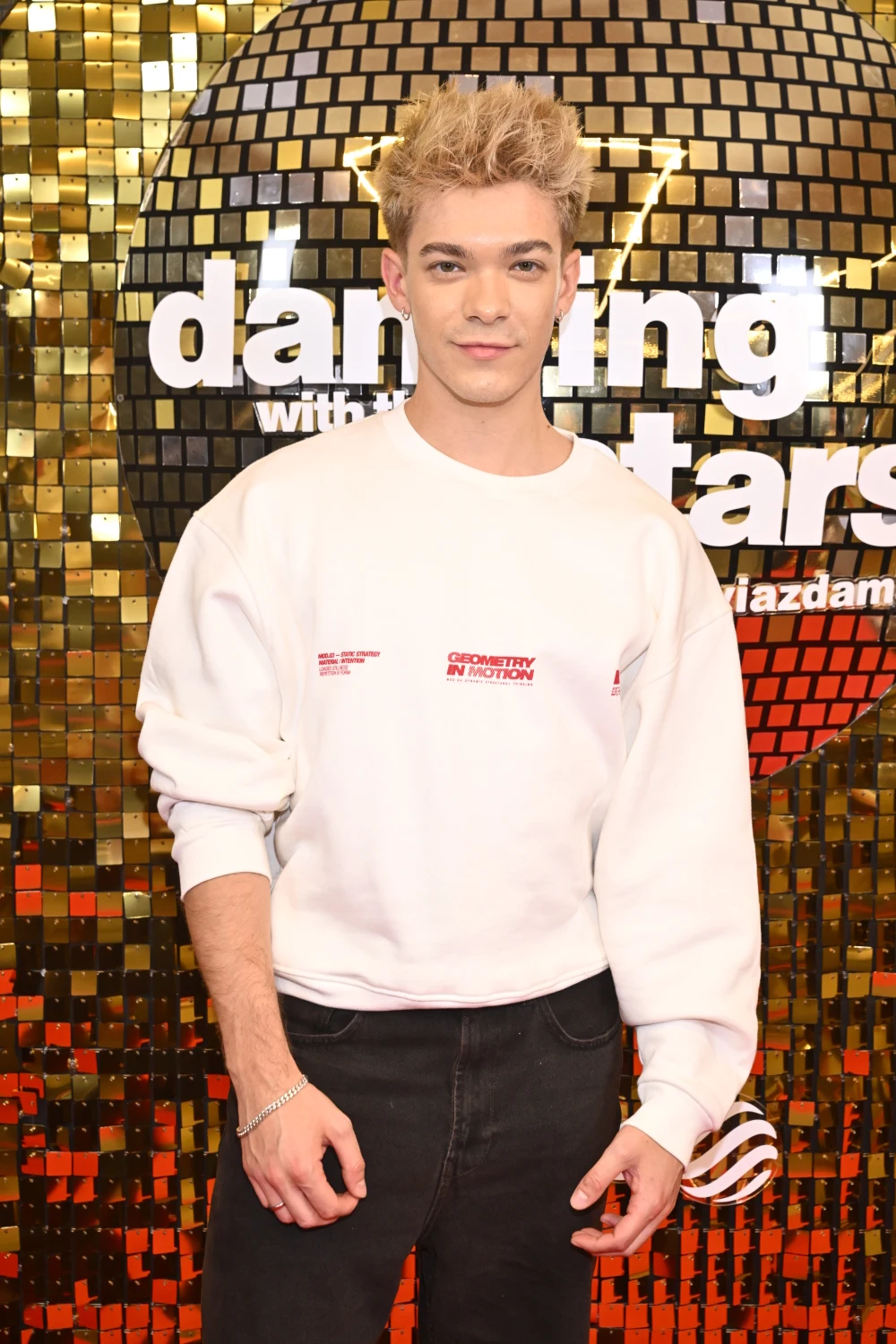 Kuba Szmajowski Młody mężczyzna z jasnymi, ułożonymi na żel włosami, ubrany w białą bluzę i czarne spodnie, pozuje na tle połyskującej złoto ściany z logo programu Dancing with the Stars.