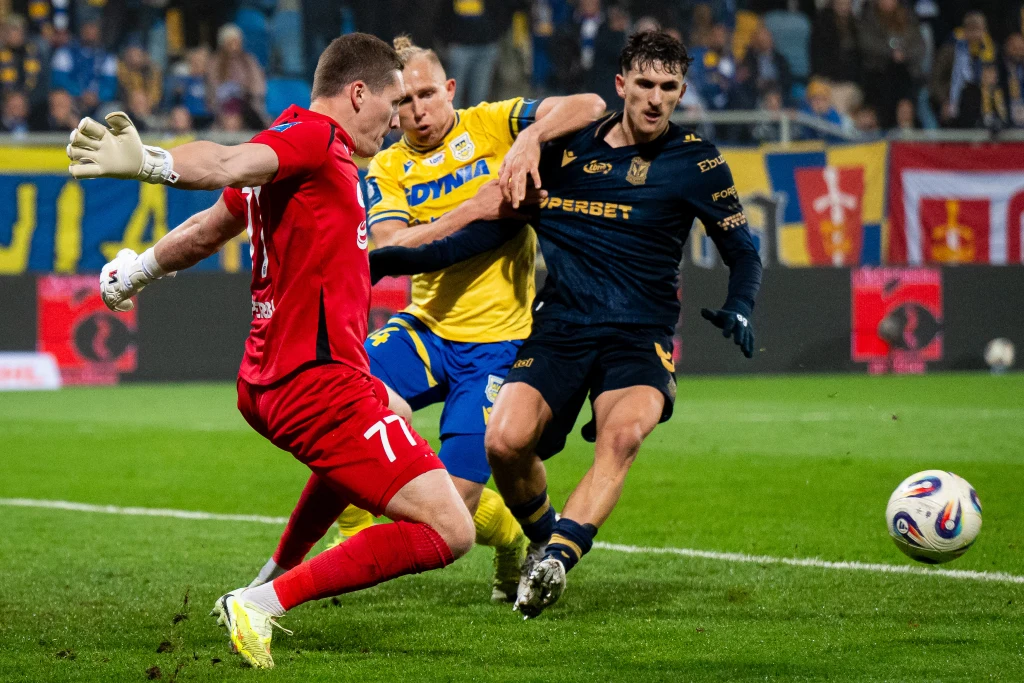 Trzech piłkarzy walczących o piłkę na tle stadionu, dwóch zawodników w ciemnych i żółtych strojach oraz bramkarz w czerwonym stroju, na tle flag kibicowskich i trybun wypełnionych widzami.