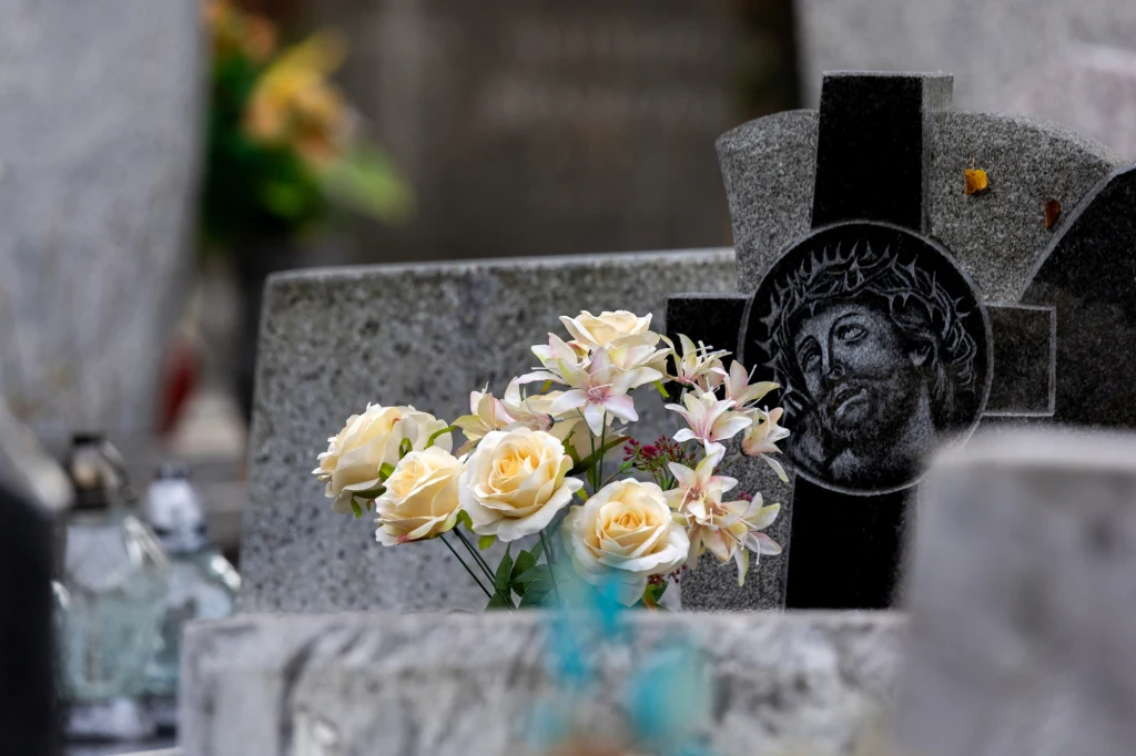 Białe i kremowe kwiaty ustawione przed nagrobkiem z czarnego granitu z wizerunkiem Jezusa Chrystusa, w tle widoczne kolejne nagrobki.