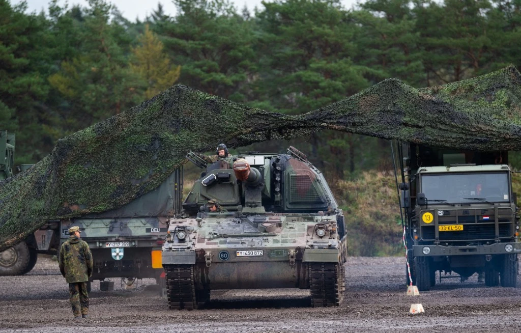 W Europie powstaje coraz więcej fabryk amunicji kalibru 155 mm (Zdj. ilustracyjne) Samosił haubica i wojskowy pojazd ciężarowy częściowo osłonięte siatką maskującą na tle leśnym, wojskowy w mundurze idący obok pojazdów, spokojna atmosfera ćwiczeń wojskowych.