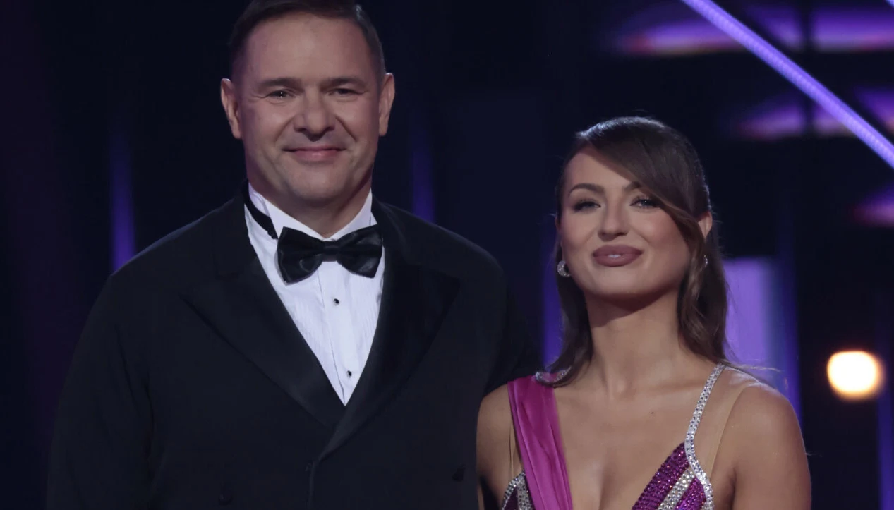 Tomasz Karolak i Izabela Skierska Elegancko ubrany mężczyzna w smokingu oraz kobieta w wieczorowej sukni stoją obok siebie na tle oświetlonej sceny.