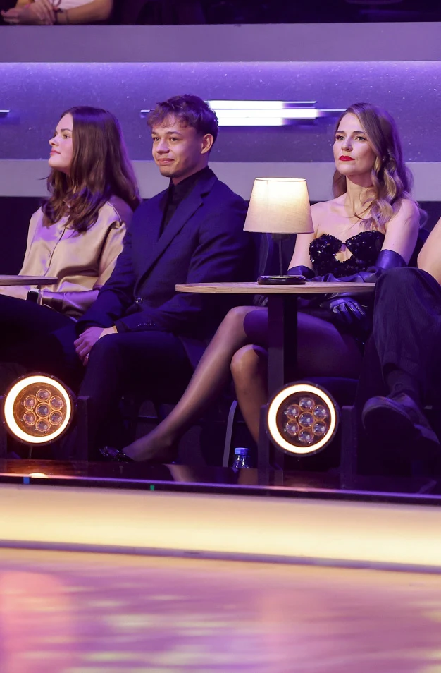 Trzy osoby siedzą przy stoliku w eleganckim wnętrzu, w tle fioletowe oświetlenie, na stoliku stoi lampka, kobieta po prawej ubrana w efektowną, czarną sukienkę z rękawiczkami, obok młody mężczyzna w garniturze, po lewej kobieta ubrana skromniej.