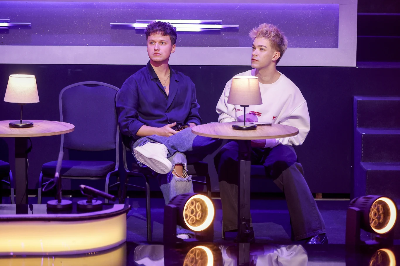 Dwóch młodych mężczyzn siedzi przy osobnych małych stolikach na scenie, z lampkami stołowymi o ciepłym świetle, otoczenie utrzymane w nowoczesnym stylu z fioletowym oświetleniem.