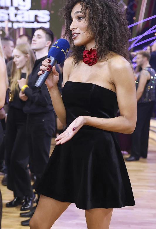 Młoda kobieta z mikrofonem ubrana w czarną, elegancką sukienkę i czerwoną ozdobę na szyi prowadzi rozmowę podczas wydarzenia. W tle widać innych uczestników oraz sceniczne oświetlenie.