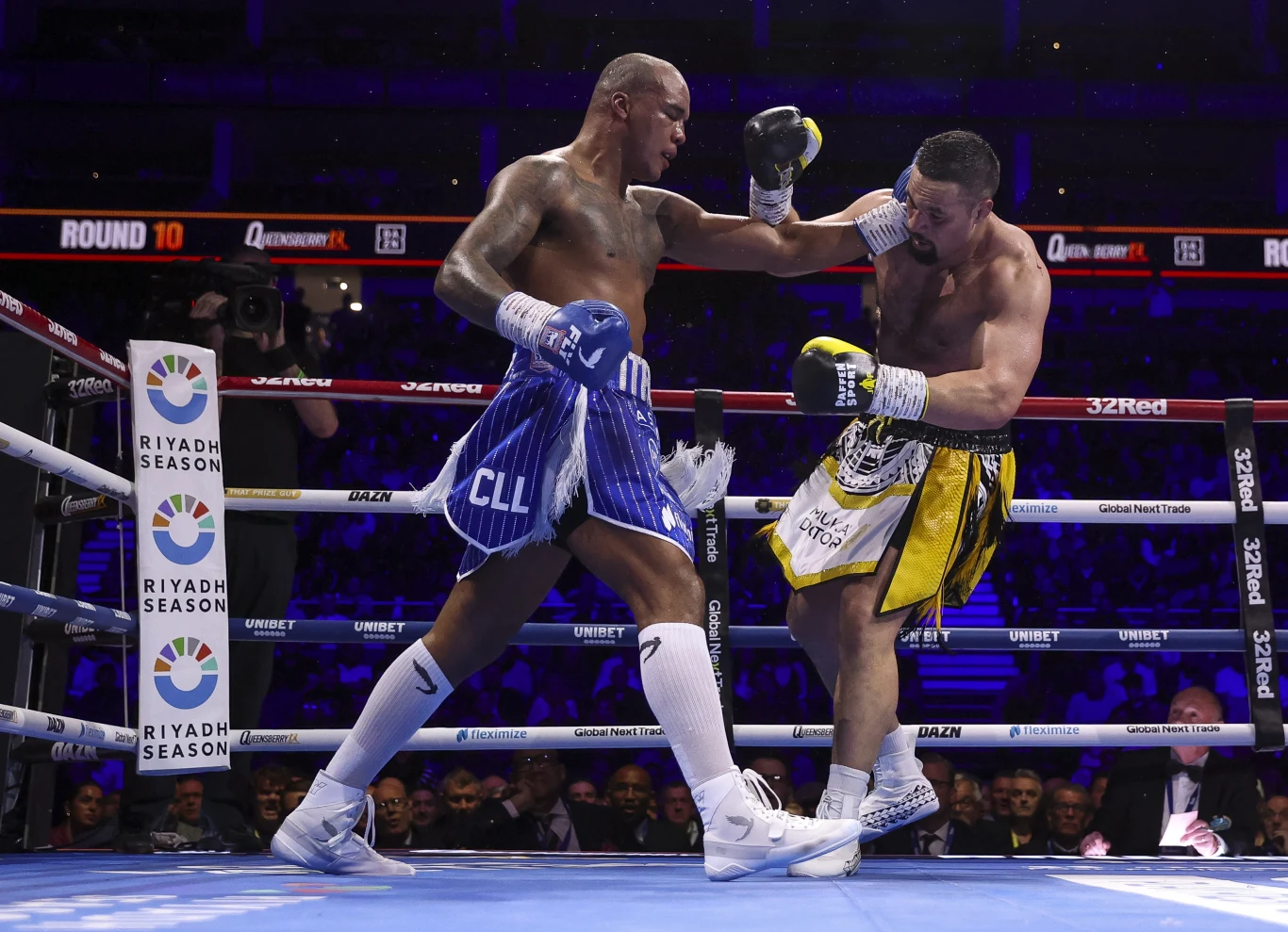 Dwóch bokserów w ringu podczas dynamicznej walki, jeden z zawodników zadaje cios drugiemu, który cofa się przed silnym uderzeniem, tło stanowią widzowie oraz niebieskie światła areny.