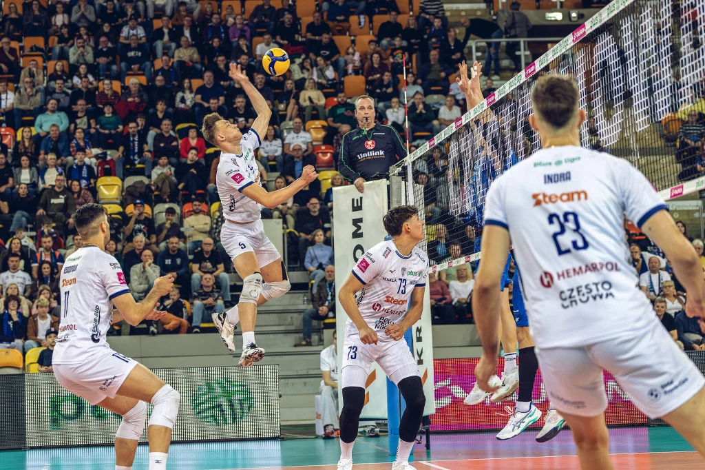 Dynamiczna akcja podczas meczu siatkówki, zawodnik drużyny w białych strojach w wyskoku odbija piłkę nad siatką, otoczony przez partnerów z drużyny i rywali po drugiej stronie boiska, a w tle wypełnione trybuny kibiców.