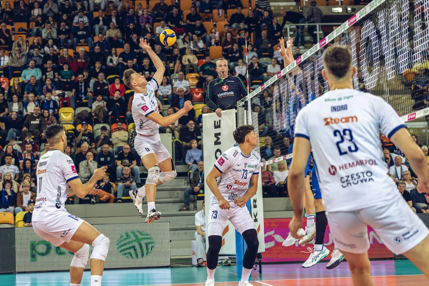 Dynamiczna akcja podczas meczu siatkówki, zawodnik drużyny w białych strojach w wyskoku odbija piłkę nad siatką, otoczony przez partnerów z drużyny i rywali po drugiej stronie boiska, a w tle wypełnione trybuny kibiców.