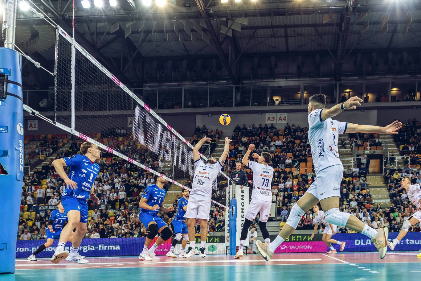 Mecz Steam Hemarpol Politechnika Częstochowa - Ślepsk Malow Suwałki Drużyna w białych strojach próbuje zablokować atak zawodnika w niebieskim stroju podczas meczu siatkówki rozgrywanego w hali sportowej pełnej kibiców. Siatka dzieli drużyny, a piłka znajduje się nad ich głowami w trakcie dynamicznej akcji.