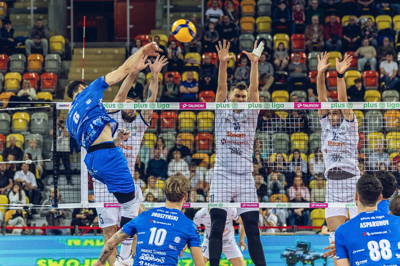 Mecz Steam Hemarpol Politechnika Częstochowa - Ślepsk Malow Suwałki Zawodnik drużyny w niebieskich strojach wykonuje atak nad siatką, trzech zawodników drużyny przeciwnej w białych koszulkach próbuje go zablokować, w tle widoczni kibice oraz trybuny.