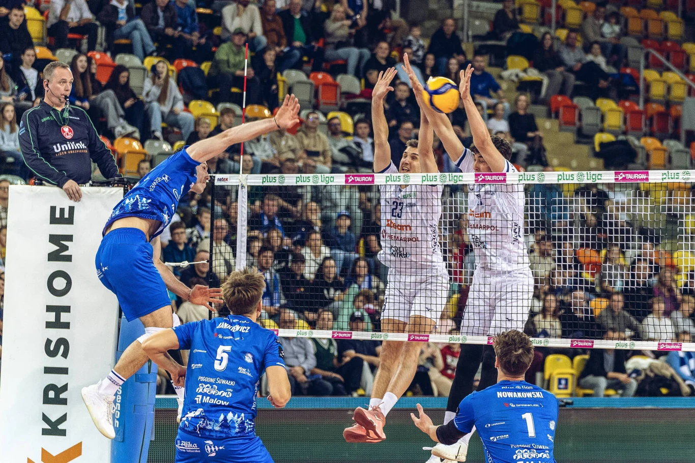 Mecz Steam Hemarpol Politechnika Częstochowa - Ślepsk Malow Suwałki Mecz siatkówki halowej, zawodnik drużyny w niebieskich strojach atakuje piłkę nad siatką, naprzeciwko dwaj gracze drużyny w białych koszulkach próbują blokować zagranie, wokół trybuny wypełnione kibicami.