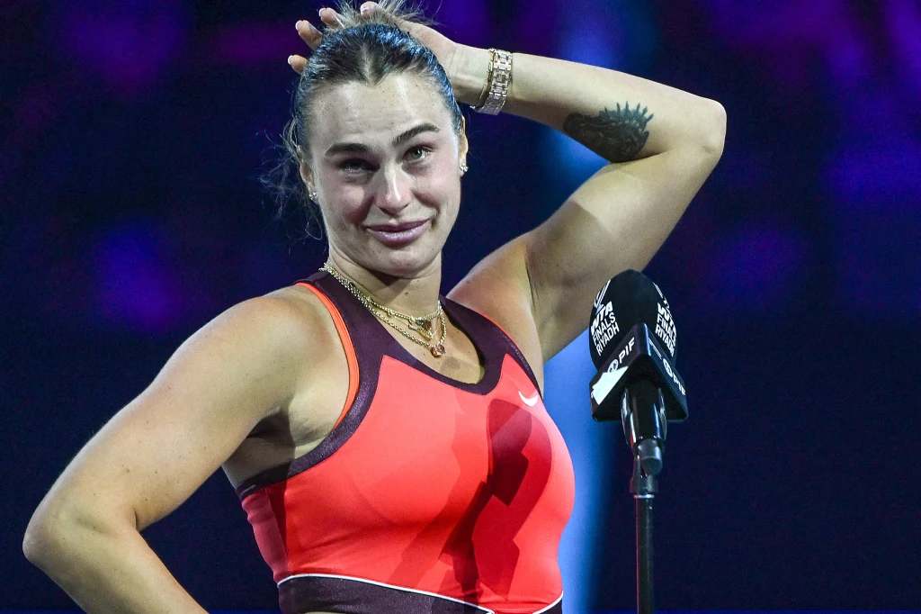 Aryna Sabalenka Zawodniczka tenisa ubrana w sportowy strój stoi przy mikrofonie na korcie, z widoczną łzą wzruszenia na twarzy i ręką uniesioną do głowy.