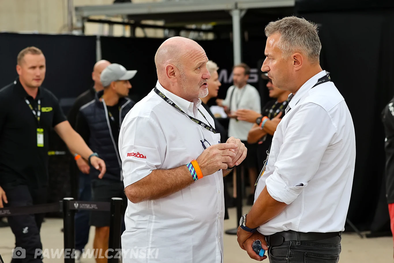 Dwóch mężczyzn w białych koszulach prowadzi rozmowę w centrum kadru, otoczeni przez innych ludzi w tle w nieformalnej atmosferze wydarzenia lub konferencji.