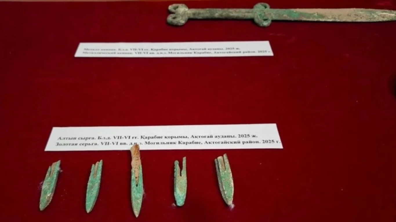 Na czerwonym tle ustawiono kilka starożytnych grotów strzał oraz miecz z wyraźnymi śladami korozji, powyżej umieszczone opisy w języku rosyjskim informują o znaleziskach archeologicznych.