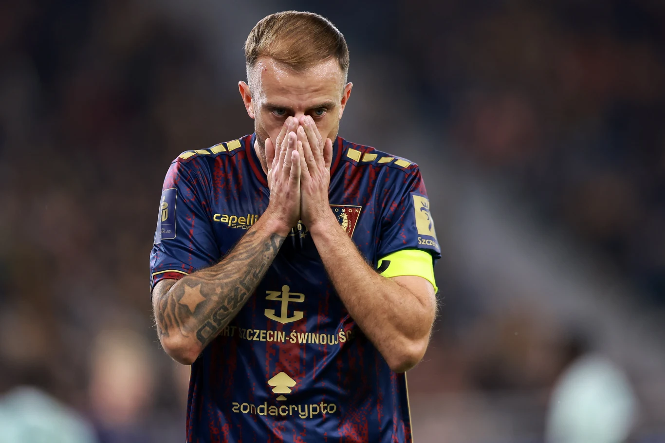 Kamil Grosicki nie kryje rozczarowania ostatnimi wynikami Pogoni Szczecin Piłkarz w stroju klubowym z opaską kapitana zasłania twarz dłońmi, wyrażając silne emocje, na tle rozmytej publiczności stadionu.