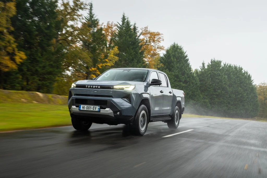 Srebrny pick-up marki Toyota podczas jazdy po mokrej nawierzchni na tle zielonych drzew i jesiennych liści, z widocznymi refleksami na asfalcie.