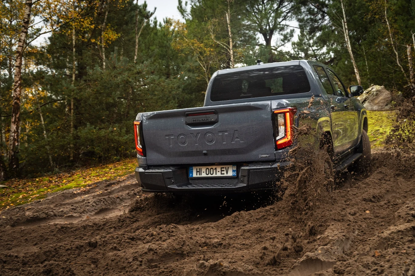 Szara Toyota jadąca off-roadowo przez błotnistą drogę, pozostawiając za sobą rozchlapaną ziemię. Samochód otoczony jest przez gęsty las z drzewami w jesiennych barwach.