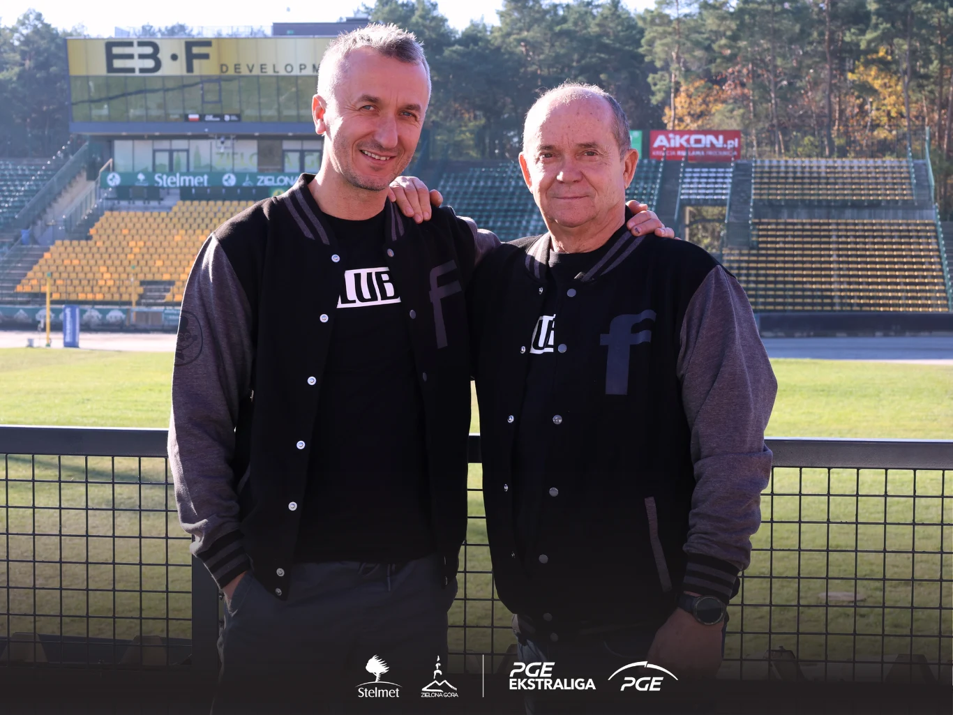 Dwóch mężczyzn stojących obok siebie na tle pustego stadionu żużlowego, obaj uśmiechnięci i ubrani w czarne bluzy z logo klubu sportowego, za nimi widoczne krzesełka na trybunach oraz logo sponsorów.